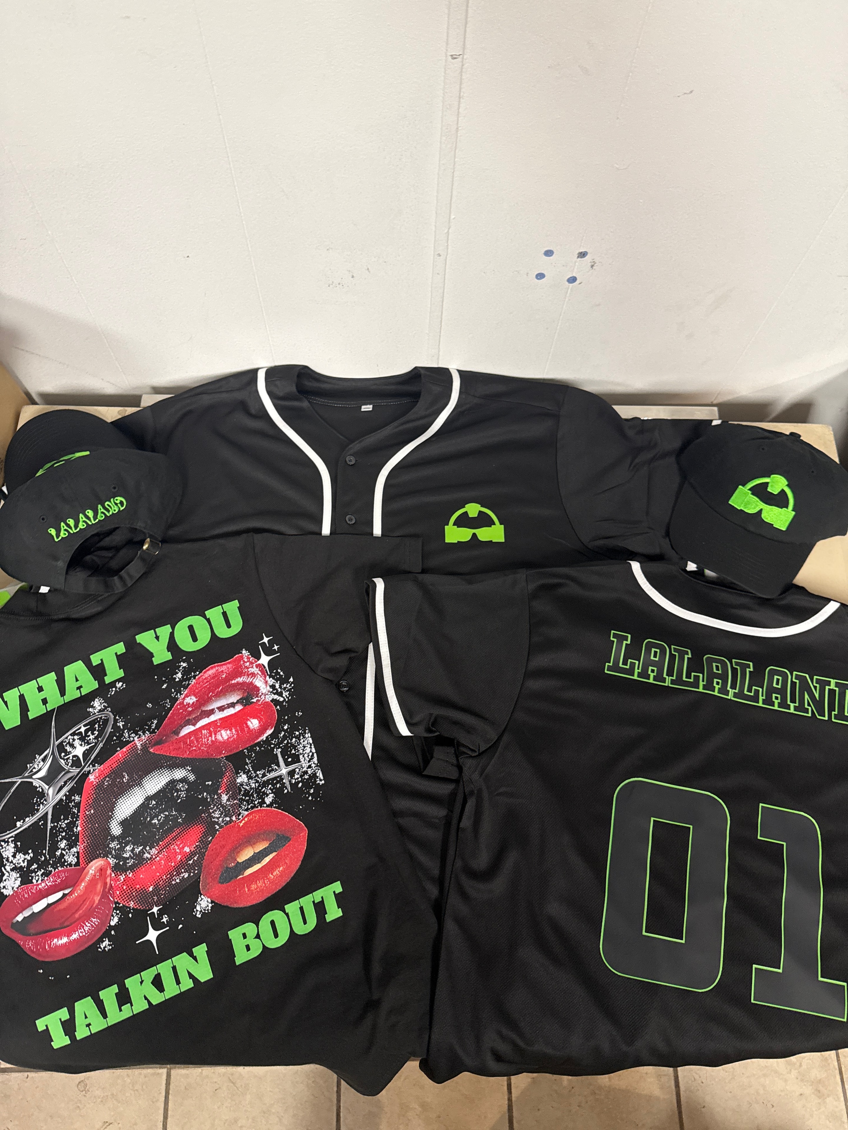 La La Land physical merch — black baseball jersey, graphic tees, and snapback caps with neon green branding